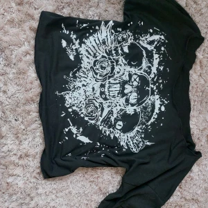 Svart off-shoulder t-shirt med döskalletryck  - Cool svart t-shirt med stort vitt tryck av döskallar och rosor på framsidan. T-shirten har korta ärmar och är off the shoulder. Bra skick, inga defekter. Pris kan diskuteras! Passar XS-M