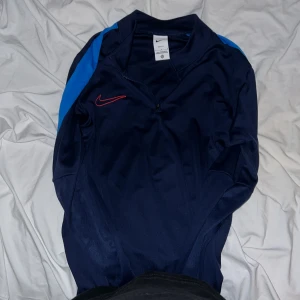 Mörkblå träningströja från Nike - Säljer en mörkblå långärmad träningströja från Nike med ljusblå detaljer på axlarna och röd Nike-logga på bröstet. Tröjan har halv dragkedja och är tillverkad i Dri-FIT material som andas. Perfekt för sport och träning.