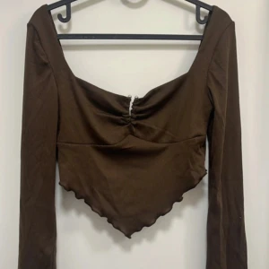 Brun långärmad topp med v-ringning - Säljer en brun långärmad topp med v-ringning och spetsig nederkant. Toppen har en snygg rynkdetalj framtill och är figurnära i modellen. Och nästan helt öppen i ryggen. Perfekt att styla med jeans eller kjol.