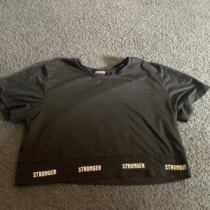 Svart croppad t-shirt från Stronger - Svart croppad t-shirt från Stronger i storlek S med texten 'STRONGER' tryckt flera gånger längs nederkanten. Den har korta ärmar och är perfekt till träning då den har ett luftigt material. Varsamt använd. 