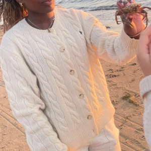 Vit kabelstickad kofta från Ralph Lauren - Säljer en klassisk vit kabelstickad kofta från Ralph Lauren med rund halsringning och knappar framtill. Koftan har långa ärmar och ribbade muddar. Perfekt för lager-på-lager och en tidlös stil.