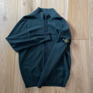 Mörkgrön ziptröja från Stone Island - Mörkgrön ziptröja från Stone Island med ribbade muddar och hög krage. Tröjan har lång ärm och klassisk logotyp-patch på vänster ärm. Perfekt för lager-på-lager och stilren look.