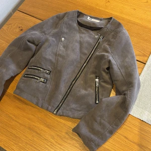 Grå mockajacka från H&M - Snygg grå mockajacka från H&M med asymmetrisk dragkedja och dekorativa blixtlåsdetaljer framtill. Jackan har lång ärm och en croppad passform som ger en cool look. Perfekt för dig som vill ha en stilren och trendig jacka.