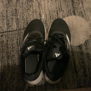 Svarta och vita sneakers från Adidas - Säljer ett par svarta och vita sneakers från Adidas med klassisk design och vit sula. Skorna har snörning och den ikoniska Adidas-loggan på plösen. Perfekta för vardagsbruk eller träning.