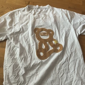 Fett T-shirt från Moschino, med en fett design på fram och baksida. - Vit t-shirt från Moschino med en stor, broderad nalleapplikation i brunt och gult på framsidan. På baksidan står det 'MOSCHINO' i matchande brodyr. Klassisk passform och rund halsringning.