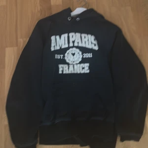 Fett hoodie från Ami Paris  - Säljer en svart hoodie från Ami Paris med vit broderad text och logga på bröstet. Tröjan har huva med dragsko och en klassisk känguruficka framtill. Perfekt för dig som gillar streetwear och vill ha något stilrent men ändå trendigt.