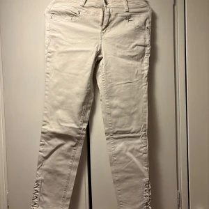 Beige byxor med snörning från Honey Blue - Snygga beige byxor från Honey Blue med unika snörningsdetaljer längst ner på benen. Byxorna har normal passform, klassiska fickor och bälteshällor. Perfekta för dig som vill ha något extra till din outfit.