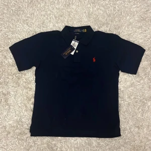 Mörkblå pikétröja från Polo Ralph Lauren - Säljer en klassisk mörkblå pikétröja från Polo Ralph Lauren med röd broderad logga på bröstet. Tröjan har korta ärmar, krage och knappar framtill. Perfekt för en stilren och sportig look.
