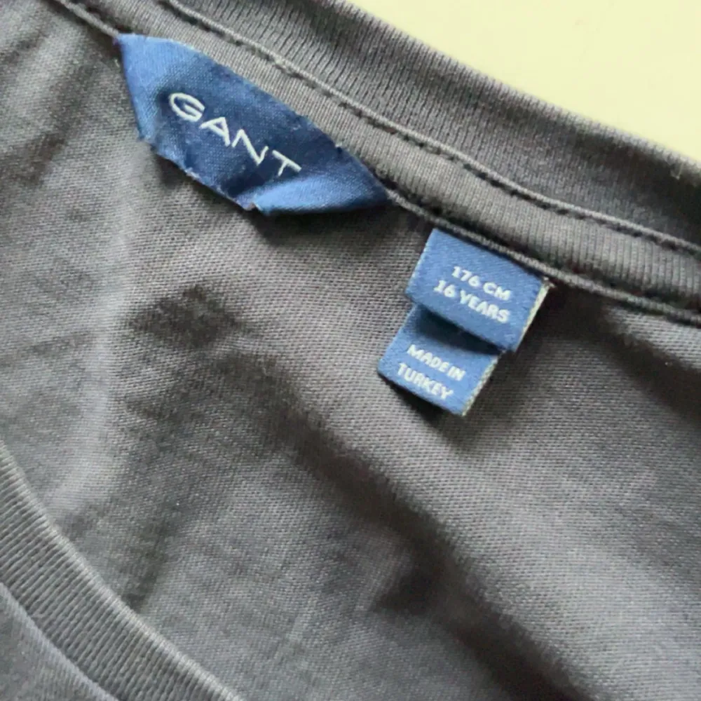 Säljer en mörkblå t-shirt från GANT med broderad logga och sköld på bröstet. Klassisk passform och rund halsringning. Perfekt för en stilren och enkel look.. T-paidat.