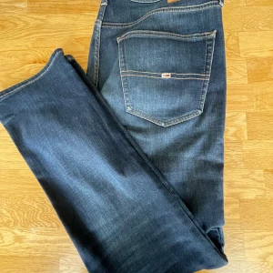 Mörkblå jeans från Tommy Jeans - Snygga mörkblå jeans från Tommy Jeans med klassisk femficksdesign och tydliga kontrastsömmar. Jeansen har en slim passform och är tillverkade i slitstarkt denim med en liten logga på bakfickan samt läderpatch i midjan. Modellen är Scantom slim