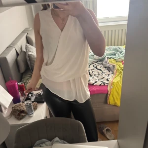 Vit omlott blus - Säljer en vit omlott blus med v-ringning och ärmlös design. Blusen har ett lager-på-lager utseende och faller snyggt över höfterna. Perfekt för dig som gillar stilrena och eleganta plagg.