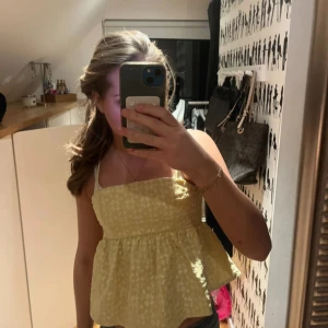 Gul blommig linnetopp med volang - Söt gul linnetopp med vita blommor och volang nertill. Toppen har smala axelband och en luftig passform, perfekt för varma dagar. Materialet känns lätt och somrigt. Den är använd ett par fåtal gånger. Tröjan är köpt utomlands. 