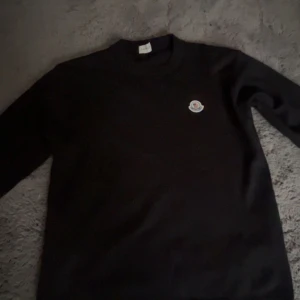 Moncler Tröja - Säljer en svart Tröja från Moncler med klassisk rund hals och Moncler-logga broderad på bröstet. Tröjan har en enkel och stilren design som passar perfekt till många olika outfits.