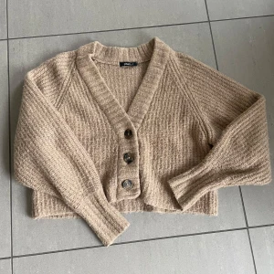 Beige stickad kofta med knappar - Mysig beige stickad kofta från Gina Tricot med v-ringning och tre stora knappar framtill. Modellen är croppad och har långa ärmar. Perfekt för lager-på-lager och en avslappnad stil.