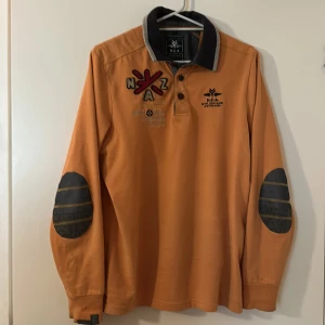 Orange långärmad pikétröja från New Zealand Auckland - Säljer en orange långärmad pikétröja från New Zealand Auckland med grå krage och knäppning framtill. Tröjan har dekorativa lappar på armbågarna och broderade detaljer på bröstet. Perfekt för en avslappnad stil. Tröjan är i storlek S. Handla via köp nu, hitta mer i profilen.