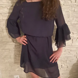 Mörkblå klänning med volangärmar - Säljer en mörkblå klänning med dubbla volanger. Klänningen har en lös passform och är tillverkad i ett lätt, transparent material med underklänning. Perfekt för dig som gillar en enkel men stilren look. Pris kan diskuteras.