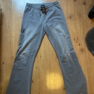 Grå mjukisbyxor från Ed Hardy - Säljer ett par grå mjukisbyxor från Ed Hardy med tryckt logga bak och coolt mönster längs ena benet. Byxorna har resår i midjan och raka ben, perfekta för en avslappnad stil.