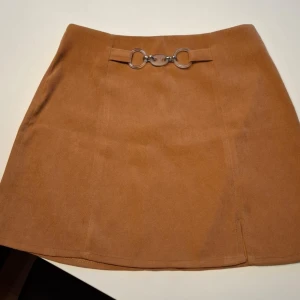  Kjol med metallspänne från Studio Mix - Säljer en kortkjol från Studio Mix i mjukt material. Kjolen har ett snyggt plastspänne framtill och dragkedja bak. Perfekt för dig som gillar stilrena detaljer och enkel design.