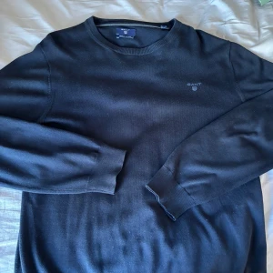 Mörk blåtröja från GANT - Klassisk mörkblå merino ull  tröja från GANT med rund halsringning och diskret logga broderad på bröstet. Perfekt för en stilren och enkel look. Tröjan har långa ärmar och ribbade muddar vid ärmslut och nederkant. Väldigt snygg tröja som passar till allt.