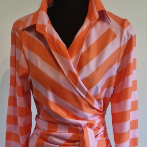 Randig omlottklänning i orange och ljusrosa - Säljer en lång omlottklänning med breda orange och ljusrosa ränder. Klänningen har lång ärm, krage och knytskärp i midjan. Snygg retrostil med markerad midja och mjukt, glansigt material.