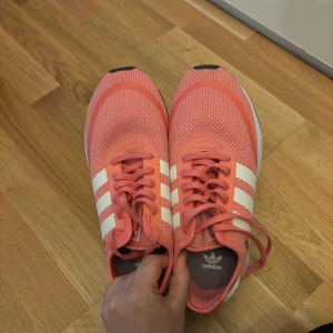 Adidas rosa sneakers med vita ränder - Säljer ett par snygga Adidas sneakers i rosa med klassiska vita ränder på sidorna. Skorna har snörning och en vit platt sula. Perfekta för dig som vill ha en färgglad och sportig look.
