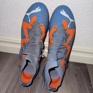 Blå och orangea fotbollsskor från Puma - Säljer ett par blå fotbollsskor från Puma med orangea detaljer och snörning. Skorna har en stickad krage och platt sula med dobbar för bra grepp på planen. Perfekta för dig som vill sticka ut på fotbollsplanen!