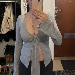 Grå långärmad knytblus från Gina Tricot  - Trendig grå långärmad topp från Gina Tricot med knyt framtill och djup v-ringning. Helt oanvänd och prislappen sitter kvar.