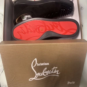 Louboutins  - Säljer ett par svarta sneakers från Christian Louboutin med ikonisk röd sula och svarta nitar på ovandelen. Skorna har en rund tå och platt sula, perfekt för dig som vill sticka ut med exklusiv design.