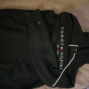 Svart hoodie från Tommy Hilfiger - Svart hoodie från Tommy Hilfiger med vit dragsko och tryckt logga samt texten 'EST. 1985' på bröstet. Klassisk huva och magficka framtill. Liten broderad flagga på ärmen. Knappt använd 