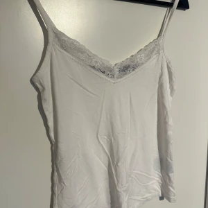 Vit linnetopp med spetskant - Säljer en vit linnetopp med smala axelband och fin spetskant längs v-ringningen. Perfekt att bära under en skjorta eller som den är under varma dagar. Enkel och stilren design. (Använd fåtal gånger, sälja pga för stor) 