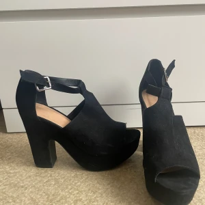 Svarta platåklackar med öppen tå - Säljer ett par svarta klackskor med platåsula och öppen tå. Skorna har en bred klack, justerbart spänne runt vristen och är tillverkade i mocka. Perfekta för dig som vill ha både höjd och stabilitet.