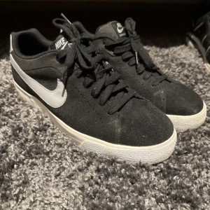 Svarta Nike sneakers med vit sula - Svarta Nike sneakers med vit sula och vit swoosh på sidan. Skorna har snörning och är tillverkade i mocka med perforerade detaljer på sidorna. Klassisk och stilren modell som passar till mycket.