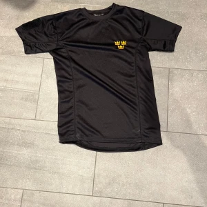 Svart t-shirt med tre kronor - Säljer en svart t-shirt med korta ärmar och tre kronor broderade i gult på bröstet. T-shirten är i ett syntetmaterial som passar perfekt till träning eller sport. Snygg och sportig stil!