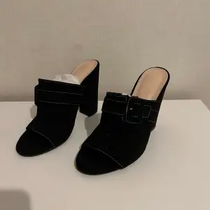 Snygga svarta klackskor med öppen tå och bred klack. Skorna har en dekorativ rem med spänne över foten som ger en stilren look. Perfekta för att lyfta din outfit till nästa nivå. Använd en gång 