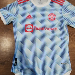 Manchester United fotbollströja från Adidas - Snygg Manchester United fotbollströja från Adidas med ett blått och vitt randigt mönster. Tröjan har korta ärmar och klubbens emblem på bröstet. Perfekt för alla fans som vill visa sitt stöd!