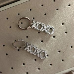 Glittriga ioaku XOXO örhängen - Snygga örhängen med glittrande XOXO-design. Perfekta för att ge en extra touch av glamour till din outfit. De är i en elegant silverfärg och har en smidig låsmekanism.