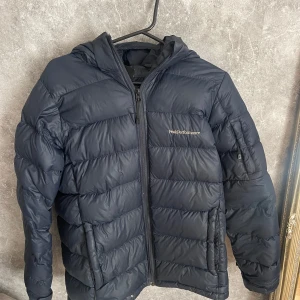 Svart dunjacka från Peak Performance - Säljer en svart dunjacka från Peak Performance. Jackan har en dragkedja framtill och är långärmad med huva. Jättefin marinblå färg, jackan är varm med två rymliga fickor framtill. Dunjackan har ett litet hål på fram sidan men som lätt går att laga. Pris går att diskutera.