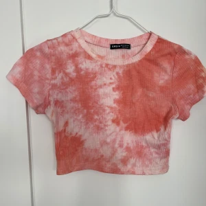 Aprikosrosa och vit croppad t-shirt från Shein - Säljer en cool croppad t-shirt från Shein i aprikosrosa och vitt batikmönster. Lite använd. En liten ljusgrå fläck på halsringningen bak (se bild) men den går förmodligen bort i tvätt. Ser ut som blyerts. 