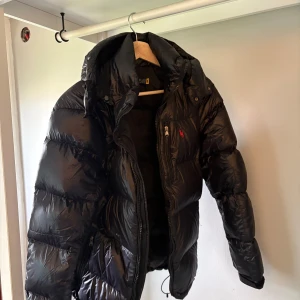 Svart dunjacka från Polo Ralph Lauren - Säljer en snygg svart dunjacka från Polo Ralph Lauren. Jackan har en pufferdesign med dragkedja och en praktisk huva. Perfekt för kyliga dagar när du vill hålla stilen.  storlek XS. Använd 3 gånger nypris 6000kr mitt pris 4000kr eller högst budande, vid snabb affär kan pris diskuteras. 