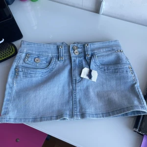 Ljusblå jeanskjol med strassdetaljer - Trendig ljusblå jeanskjol med snygga strassdetaljer på fickorna och ett dekorativt bälte. Kjolen har en kort längd och är perfekt för en cool och avslappnad stil. Den har också knappar och dragkedja framtill.