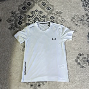 Vit t-shirt från Under Armour - Säljer en vit t-shirt från Under Armour med korta ärmar och logga på bröstet. Perfekt för träning eller en avslappnad dag. T-shirten har en sportig look med text på sidan. Helt ny oanvänd 