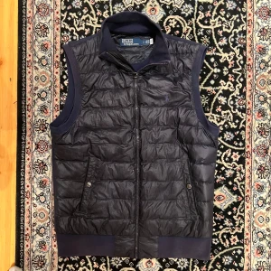 Mörkblå dunväst från Polo Ralph Lauren - Säljer en snygg mörkblå dunväst från Polo Ralph Lauren. Västen har en dragkedja framtill och två fickor med knappar. Perfekt för lager-på-lager under kyliga dagar. Klassisk och stilren design. Super fin i skick endast några få skador som knappt syns vid användning 