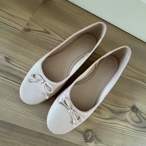 Ljusrosa ballerinaskor med rosett - Säljer ett par eleganta ljusrosa ballerinaskor med en söt rosett på ovansidan. Skorna har en rund tå och är tillverkade i ett syntetmaterial med en diskret textur. Perfekta för en stilren look. Endast använda en gång. 