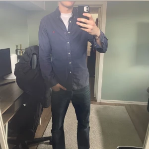 Mörkblå skjorta från Ralph Lauren - Säljer en stilren mörkblå skjorta från Ralph Lauren i slim fit. Skjortan har långa ärmar och knappar framtill. Den är tillverkad i ett bekvämt material och har en liten röd broderad logga på bröstet. Perfekt för en snygg och avslappnad look.