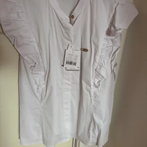 Vit blus från U.S. Polo Assn. - Säljer en stilren vit blus från U.S. Polo Assn. med volangdetaljer längs ärmarna och en v-ringad design. Blusen har en slim passform och knappar framtill. Perfekt för en elegant look. (aldrig använt)