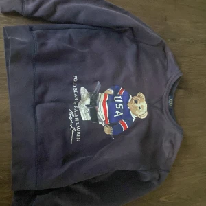 Mörkblå tröja med Polo Bear från Ralph Lauren - Säljer en mörkblå tröja från Ralph Lauren med den ikoniska Polo Bear i en USA-tröja. Tröjan har långa ärmar och är perfekt för en avslappnad stil. Den är gjord i ett mjukt material som känns skönt mot huden.