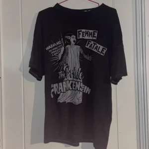 Svart t-shirt med Bride of Frankenstein-tryck från H&M Divided - Cool svart t-shirt från H&M Divided med ett stort tryck av Bride of Frankenstein och texten 'Femme Fatale'. Perfekt för dig som gillar klassiska skräckfilmer och vill ha en unik stil. T-shirten har korta ärmar och en normal passform.