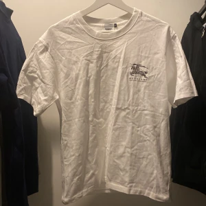 Vit och svart t-shirt från Burberry - Säljer två stilrena t-shirts från Burberry i storlek S. En vit och en svart, båda med ett diskret tryck på bröstet. Perfekta för en casual look. Passa på att köpa dessa klassiska plagg!