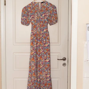 Blommig klänning med öppen rygg - Säljer en färgglad blommig klänning med korta ärmar och en snygg öppen ryggdetalj. Klänningen har ett knytband i midjan och en knapp i nacken. Använd ett fåtal gånger men har inga tecken på användning. Perfekt för sommarens alla tillfällen!