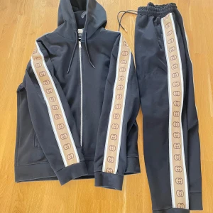 Svart hoodie och byxor från Gucci - Jag är 1.90cm väger 85kg sitter lite stort på mig…. Säljer en snygg svart hoodie och matchande byxor från Gucci. Setet har beige och bruna detaljer längs ärmar och ben med det ikoniska Gucci-mönstret. Hoodien har dragkedja och justerbar huva. Perfekt för en stilren och bekväm look.